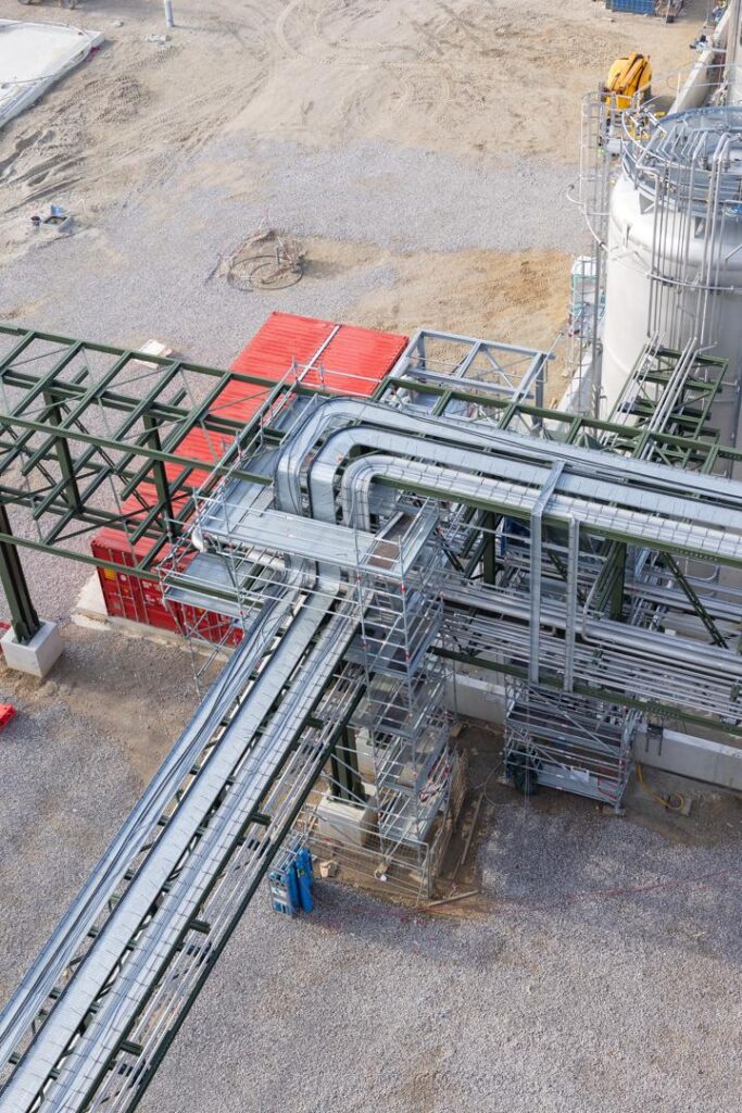 Vue aérienne plongeante montrant l'imbrication complexe d'un échafaudage industriel autour d'un réseau de tuyauterie (pipe rack) et de chemins de câbles pour la maintenance